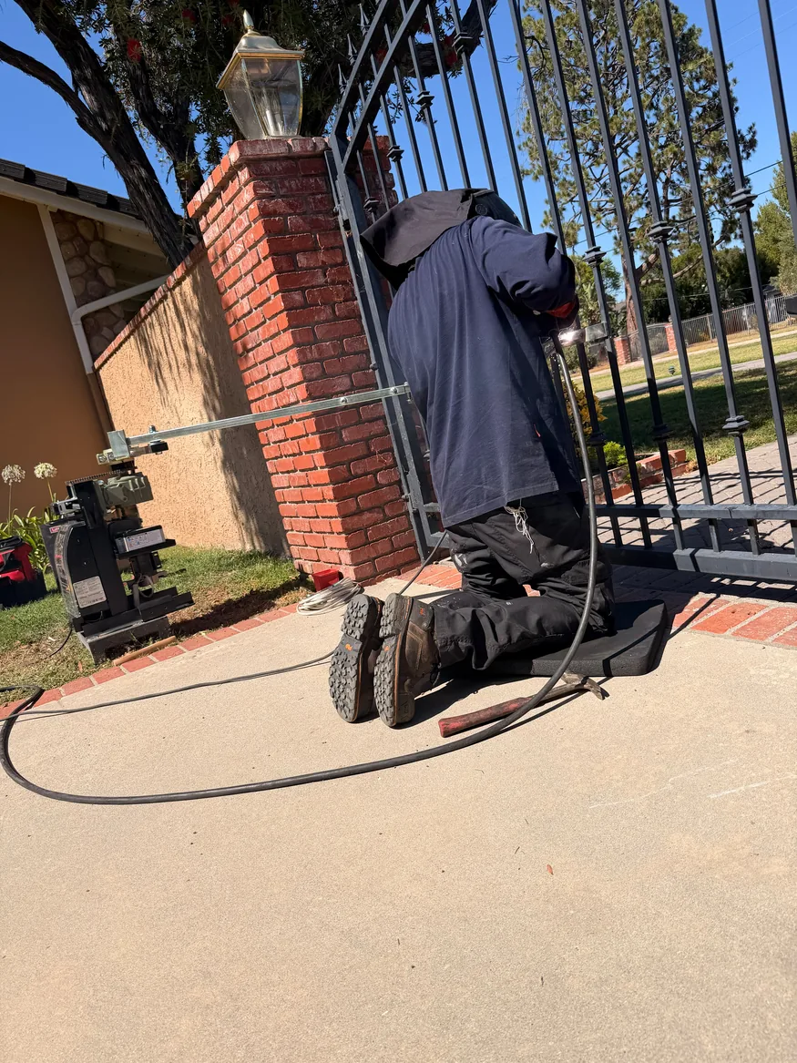 Technician welding a metal driveway gate attachment beside a brick entrance pillar