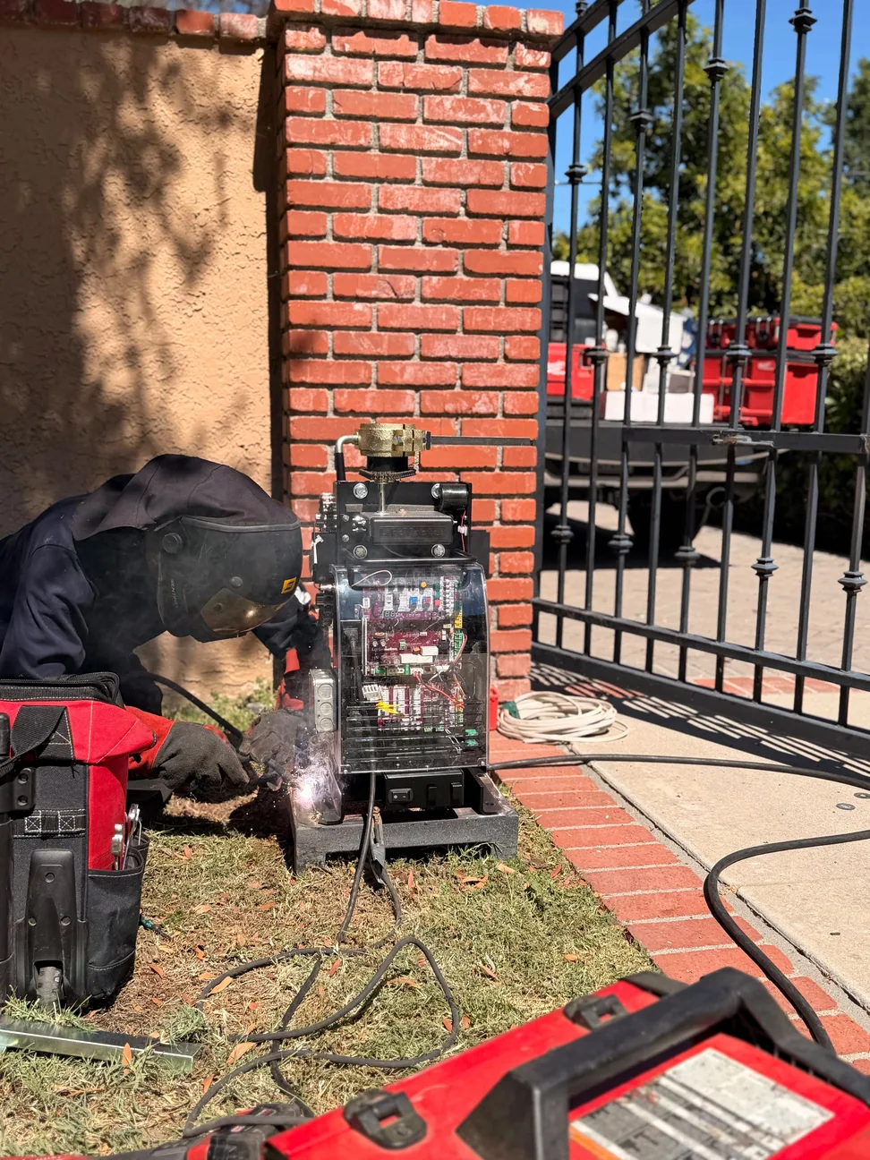 Technician welding beside an exposed gate operator at a metal driveway gate and brick pillar