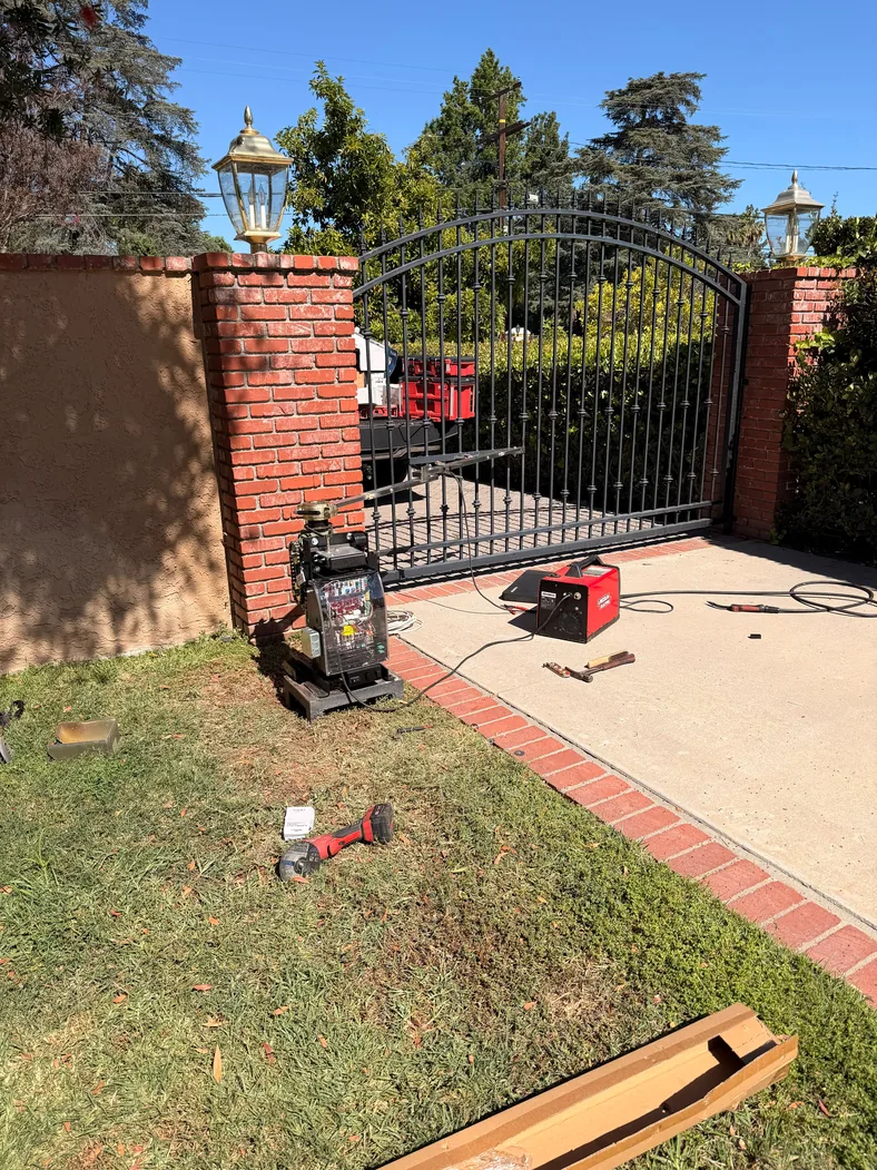 Arched metal driveway gate between brick pillars with an exposed operator and welding equipment